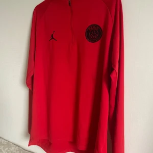 Jordan x PSG sweatshirt  - Säljer en snygg röd långärmad tröja från Jordan x PSG. Tröjan har en halv dragkedja och det ikoniska PSG-emblemet på bröstet. Perfekt för fotbollsfans som vill ha något stilrent och bekvämt. Passar bra till både träning och vardag.