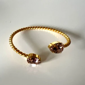 Guldarmband med stenar från Caroline Svedbom - Snyggt guldarmband med en tvinnad design och två stora lila stenar i ändarna. Armbandet är öppet och justerbart för att passa de flesta handleder. Perfekt för festliga tillfällen eller som en elegant vardagsaccessoar.. Använd vid ett tillfälle! Färgen Vintage Rose på stenen