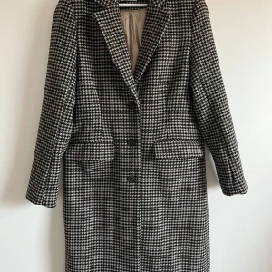 Rutig kappa - Säljer en superfin rutig kappa i svart och beige. Den har en klassisk design med knappar framtill och långa ärmar. Perfekt för höst och vår, och ger en stilren look till vilken outfit som helst. Passar både till vardags och lite finare tillfällen.