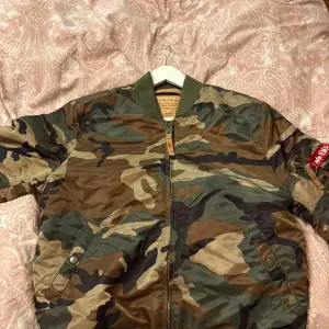 Säljer en snygg camouflage bomberjacka från Alpha Industries i storlek L. Jackan har en klassisk camo-look med dragkedja och orange foder. Perfekt för höst och vår, och den har en cool detalj med en röd tagg på ärmen. Passar både till vardags och lite mer avslappnade tillfällen.