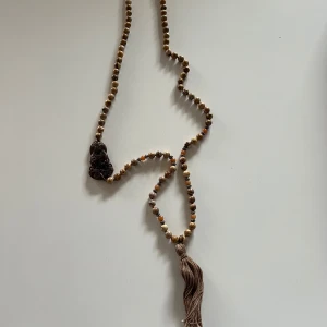 Yoga halsband med tofs från soul of Maia  - Unikt bohemiskt halsband med pärlor i jordnära toner och en stor tofs i beige. Halsbandet har en mörk dekorativ detalj som ger en mystisk touch. Perfekt för att ge din outfit en avslappnad och naturlig stil.