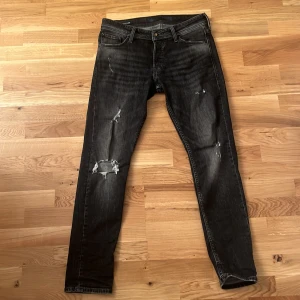 Svarta slitna jeans från Jack & Jones - Säljer ett par svarta jeans från Jack & Jones i slim fit. De har en cool sliten look med flera revor och slitningar. Perfekta för en avslappnad stil. Midjan är midwaist och de har klassiska fem fickor. Passar perfekt till både vardag och fest!
