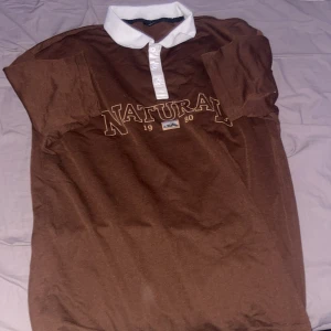Brun pikétröja med texten 'NATURAL' - Säljer en brun pikétröja med vit krage och knappar. På framsidan står det 'NATURAL 1980' i stora bokstäver. Perfekt för en avslappnad stil eller vardagsbruk. Tröjan är i bra skick och redo för nya äventyr!