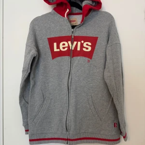 Grå hoodie från Levi's strl: 16 år - Säljer en snygg grå hoodie från Levi's med röd insida i huvan och stort rött Levi's-tryck på bröstet. Den har en dragkedja framtill och praktiska fickor. Perfekt för en avslappnad stil och passar bra till både höst och vår.