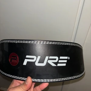 Svart träningsbälte från Pure - Säljer ett svart träningsbälte från Pure i mycket bra skick. Bältet är gjort av skinn och har en vadderad insida för extra komfort. Det har vita sömmar och ett justerbart spänne med flera hål för perfekt passform. Perfekt för styrketräning och gympass!