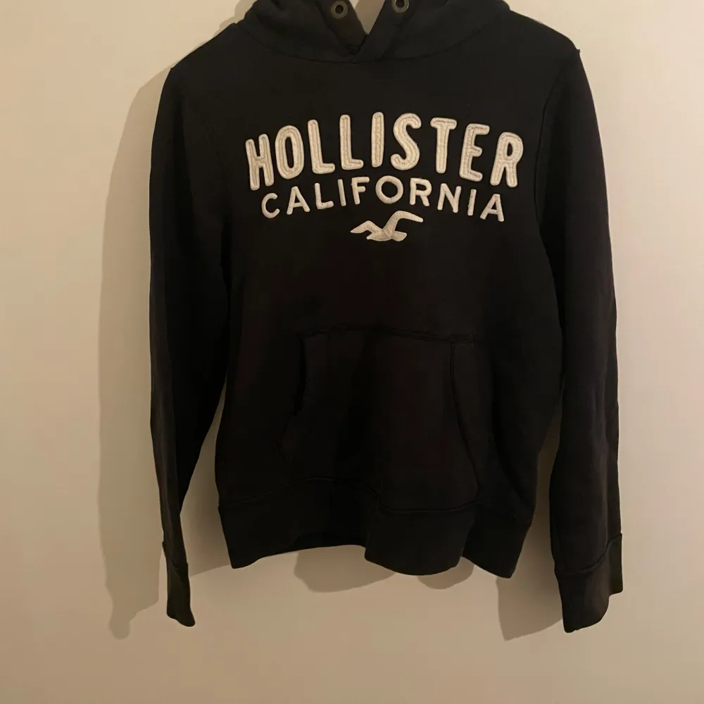 Säljer en svart hoodie från Hollister med texten 'Hollister California' i vitt på framsidan. Den har en klassisk känguruficka och en bekväm huva med dragsko. Perfekt för en avslappnad stil eller kyliga dagar. Passar både till vardags och mysiga hemmakvällar.. Tröjor & Koftor.