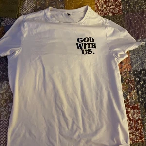 Vit t-shirt med tryck - Säljer en vit t-shirt med texten 'GOD WITH US.' tryckt på både fram- och baksidan. På baksidan finns även en jordglob i blått och texten 'WATER COVERS A LARGE PROPORTION OF THE EARTH'S SURFACE.' T-shirten är kortärmad och i bomull, den är oversized 