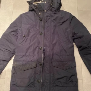 Mörkblå parkajacka från J.Lindeberg - Säljer en snygg mörkblå parkajacka från J.Lindeberg i storlek S. Jackan har en avtagbar huva med pälsdetaljer och stängs med både dragkedja och knappar. Nypris ca 4000kr. Använd en vinter. Perfekt för kalla vinterdagar! 🧥❄️