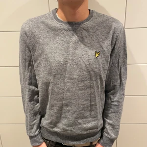 Grå tröja från Lyle & Scott - Säljer en stilren grå tröja från Lyle & Scott i mycket bra skick. Den har en klassisk rund hals och ett broderat gult emblem på bröstet. Perfekt för både vardag och lite finare tillfällen. Passar bra till jeans eller chinos! Är i storlek L men sitter bra till både L och M!!! Pris 70kr  , MVH Liam 