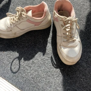 Vita sneakers med rosa detaljer från My Shoes - Säljer ett par vita sneakers med rosa detaljer från NLY Shoes i storlek EU 37. Skorna har snörning och är i acceptabelt skick med lite slitage på insidan. Perfekta för vardagsbruk och ger en söt touch till din outfit!