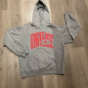 Grå hoodie från Vailent - Säljer en grå hoodie från Vailent i storlek L. Den har en stor röd text 'UNIVERSE' på framsidan och en bekväm passform. Perfekt för höst och vinter med sin mysiga huva och långa ärmar. Använd fåtal gånger
