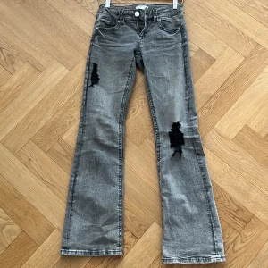 Grå bootcut jeans med slitningar storlek 158 - Säljer ett par grå bootcut jeans med slitna detaljer på båda benen. Jeansen har klassisk femficksdesign, knäppning med dragkedja och knapp, samt vida ben nertill. Perfekta för en avslappnad och trendig look.