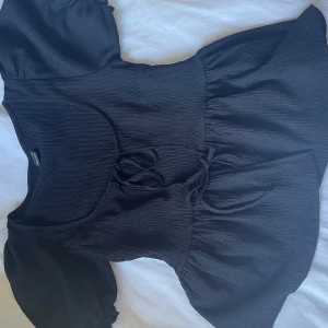 Svart ribbad topp från Gina Tricot - Svart ribbad topp från Gina Tricot med korta puffärmar och knytning framtill i midjan. Toppen har en figurnära passform upptill och volang nedtill för en söt och trendig look.