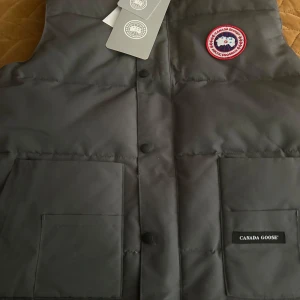 Svart dunväst från Canada Goose - Säljer en svart dunväst från Canada Goose i färgen grå och med röd logga.     Storlek xl men passar både m och L 