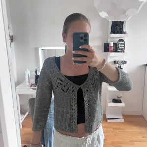 Grå glittrig kofta från KappAhl - Säljer en grå glittrig kofta från KappAhl med fina hålstickade detaljer och trekvartsärm. Koftan är öppen framtill och har en lätt croppad passform. Perfekt att slänga över ett linne eller klänning för en extra touch.