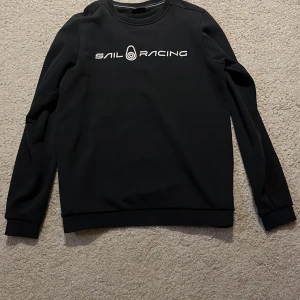 Svart sweatshirt från Sail Racing - Svart sweatshirt från Sail Racing med vit logotyp på bröstet. Tröjan har rund halsringning och långa ärmar. Mjuk insida och ribbade muddar vid ärmslut och nederkant. Perfekt för en stilren och sportig look.
