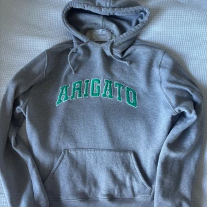 Grå hoodie från Axel Arigato - Grå hoodie från Axel Arigato med grön text 'ARIGATO' på bröstet. Storlek S.