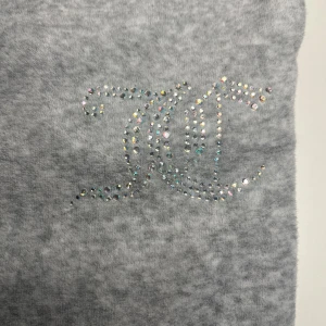 Grå mjukisbyxor med strasslogga från Juicy Couture - Säljer ett par grå mjukisbyxor från Juicy Couture med deras logga i glittrande strass på ena benet. Byxorna har en mjuk och skön känsla, perfekta för en avslappnad stil. Resår i midjan för extra komfort. Jag säljer pga av att de har blivit för små. Priset kan diskuteras💗