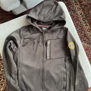 Svart hoodiejacka från Zavetti Canada  reflektion - Svart hoodiejacka med dragkedja från Zavetti Canada. Jackan har en stor lönnlövsdetalj på ryggen, bröstficka med dragkedja och patch på ärmen. Perfekt för dig som gillar stilren design med sportig känsla. Stl: M barn, passar 10-11 åringar.