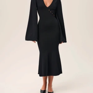 Svart långklänning med v-ringning och vida ärmar - Elegant svart långklänning med djup v-ringning och dekorativa knappar framtill. Klänningen har långa, vida ärmar och figurnära passform som framhäver siluetten. Perfekt för dig som vill ha en stilren och sofistikerad look.