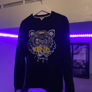 Svart sweatshirt med tigertryck från Kenzo - Svart sweatshirt från Kenzo med ikoniskt tigerbroderi och texten 'KENZO PARIS' framtill. Tröjan har rund halsringning och långa ärmar. Perfekt statement-plagg för dig som gillar streetwear och snygga detaljer.