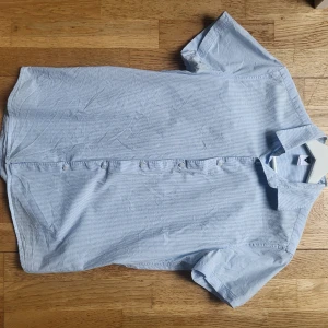 Ljusblå randig skjorta i nytt skick - Säljer en ljusblå kortärmad skjorta i nytt skick. Skjortan har knäppning framtill och är i ett lätt och svalt material, perfekt för varmare dagar. Den bör passa ca 165-185 beroende på hur tight man vill att den ska sitta. Priset är inte satt i sten, så kontakta gärna!