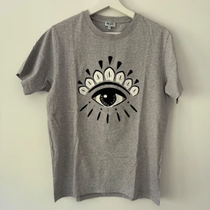 Grå t-shirt med ögonmotiv från Kenzo - Säljer en grå t-shirt från Kenzo med ett stort svartvitt ögonmotiv på bröstet. T-shirten har rund halsringning och korta ärmar. Perfekt för dig som gillar unika prints och streetwear vibes.