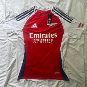 Arsenal hemmatröja 2023/24 från Adidas - Säljer en Arsenal hemmatröja från Adidas med röd framsida, vita ärmar och blå detaljer. Tröjan har klubbmärke och sponsortryck på bröstet samt Adidas-logga. Klassisk design med rund hals och korta ärmar. Säljer tröjan billigt för att trycket är sne 