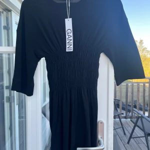 Svart klänning från Ganni med smock i midjan - Svart klänning från Ganni med smockad midja och halvlånga ärmar. Klänningen har en rund halsringning och är tillverkad i ett mjukt, stretchigt material. Perfekt för dig som gillar stilrena och bekväma plagg.