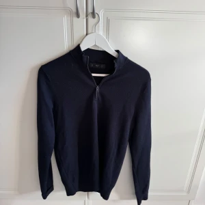 Mörkblå stickad tröja med dragkedja från Mango Man - Säljer en mörkblå stickad tröja från Mango Man i storlek M. Tröjan har halv dragkedja framtill och lång ärm, perfekt för lager-på-lager. Enkel och stilren design som passar till mycket.