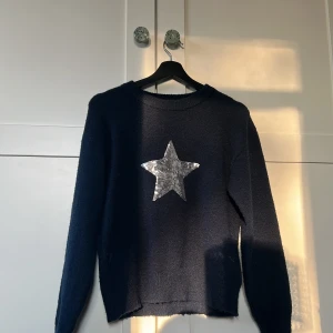 Mörkblå stickad tröja med paljettstjärna från Gina Tricot - Mysig mörkblå stickad tröja från Gina Tricot med en stor silverfärgad paljettstjärna på bröstet. Tröjan har rund halsringning och långa ärmar. Perfekt för dig som vill sticka ut lite extra!