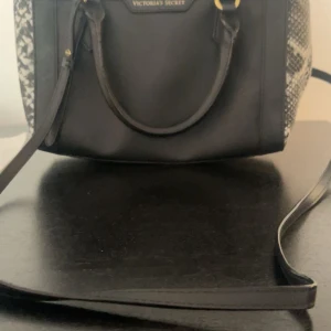 Svart handväska med ormmönster från Victoria's Secret - Snygg svart handväska från Victoria's Secret med ormmönstrade sidor och guldfärgade detaljer. Väskan har både handtag och axelrem samt dragkedja upptill. Perfekt för att lyfta din outfit!