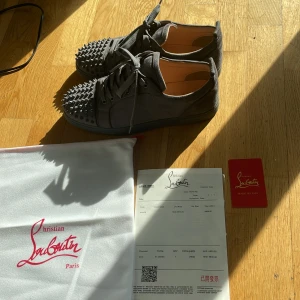 Christian Louboutin (loubs) - Säljer ett par gråa sneakers från Christian Louboutin i mocka med ikoniska röda sulor. Skorna har snörning och är dekorerade med nitar framtill för en edgy look. Perfekta för dig som vill sticka ut med exklusiva detaljer🌟Skriv till mig vid minsta fundering💫