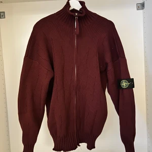 Vinröd stickad ziptröja från Stone Island - Vinröd stickad tröja från Stone Island med hel dragkedja framtill och hög krage. Tröjan har långa ärmar och ribbade muddar samt den klassiska Stone Island-loggan på ärmen. Perfekt för lager-på-lager-stil.