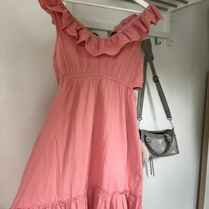 Rosa volangklänning med broderad krage - Supersöt rosa klänning med volangdetaljer och broderad krage. Ärmlös modell med avslappnad passform och lätt rynk vid midjan. Perfekt för varma dagar och somriga tillfällen.