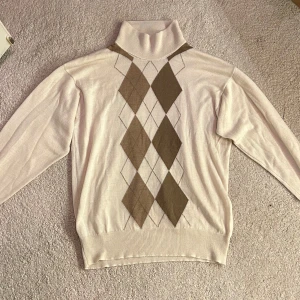 Beige stickad polotröja med argylemönster - Säljer en beige stickad polotröja med klassiskt argylemönster i bruna och ljusbruna toner framtill. Tröjan har långa ärmar och ribbade muddar. Perfekt för dig som gillar en preppy och stilren look.