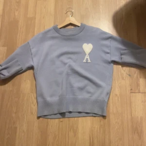Ljuslila stickad tröja med hjärtdetalj - Säljer en ljus blå Ami paris som är  stickad tröja med ribbade muddar och en vit hjärtapplikation med bokstaven A på bröstet. Tröjan har lång ärm och en avslappnad passform, perfekt för kyliga dagar.priset kan diskuteras 