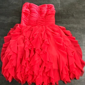 Röd volangklänning med axelbandslös design - Säljer en fin axelbandslös röd klänning med volanger. Köpt här på Plick, men hittar ingen lapp vart den är från. Bra skick! Säljer då den är för liten på mig!