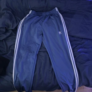 Blå Adidas träningsbyxor med vita ränder - Säljer ett par klassiska blå träningsbyxor från Adidas med vita ränder längs sidorna och dragkedjor vid fickorna. Byxorna har resår i midjan och är perfekta för sport eller chill.