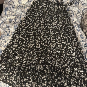 Svart blommig långklänning - Säljer en svart långklänning med vitt blommigt mönster. Klänningen har en lätt utsvängd form och volangdetalj nertill. Perfekt för dig som gillar en flowy och feminin stil.