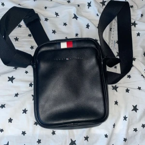 Svart axelväska från Tommy Hilfiger - Snygg svart axelväska från Tommy Hilfiger med justerbart band och klassiska Hilfiger-färger på framsidan. Väskan har dragkedja och är tillverkad i skinnimitation. Perfekt för att bära det viktigaste med stil.
