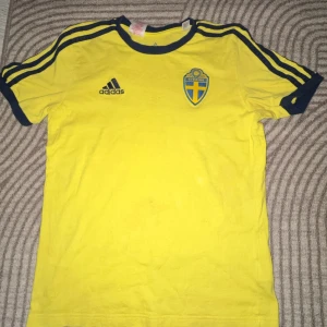Svenska landslagets fotbollströja från Adidas - Gul kortärmad fotbollströja från Adidas med blå detaljer och Sveriges landslagsemblem på bröstet. Klassisk design med blå ränder på axlarna och mörkblå Adidas-logga. Perfekt för dig som vill visa ditt stöd för Sverige. Har två små hål men annars top skick 🤩