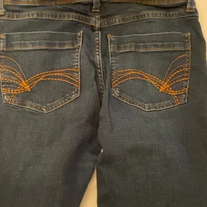 Blå jeansbyxor från Divided med orange sömmar - Säljer ett par klassiska blå jeansbyxor från Divided med snygga orange dekorationssömmar på bakfickorna. Byxorna har normal passform och raka ben. Perfekta till många olika stilar!