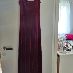 Vinröd sammetsklänning från Adelsten - Säljer en vinröd långklänning i sammetsliknande material från Adelsten. Klänningen har smala axelband och en rak, elegant siluett. Perfekt för dig som vill sticka ut med en lyxig känsla och djup färgton.