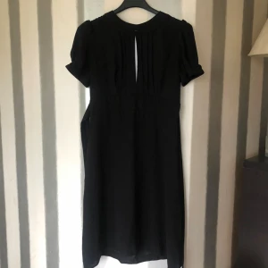 Svart klänning med kort ärm och knappdetalj - Svart klänning med korta puffärmar och knappdetalj vid ärmslut. Klänningen har en liten öppning framtill vid halsen och en större öppning i ryggen med knappar. Stilren och enkel design med markerad midja. Storlek är 34/36