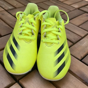 Gula Adidas fotbollsskor med svarta ränder - Säljer ett par knallgula Adidas fotbollsskor med klassiska svarta ränder på sidorna. Skorna har snörning och är tillverkade i syntetmaterial. Perfekta för dig som vill sticka ut på planen!