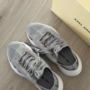 Grå Axel Arigato sneakers - Säljer ett par helt nya gråa Apex Flash runner Axel Arigato sneakers i storlek 36. Skorna har snörning och diskreta svarta detaljer i mönstret. Skokartongen medföljer! 