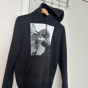 Svart hoodie med Tupac-tryck - Svart hoodie med stor svartvit bild av Tupac på bröstet. Tröjan har huva och en stor magficka framtill. Perfekt för dig som gillar streetwear och vill ha en bekväm och snygg tröja med ikoniskt tryck.