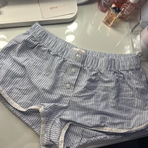 Blåvitrandiga shorts med spets från Gina Tricot - Söta blåvitrandiga shorts från Gina Tricot med vita knappar framtill och elastisk midja. Dekorerade med vit spets och en liten rosett i sidan. Perfekta för varma dagar eller som mysplagg hemma.
