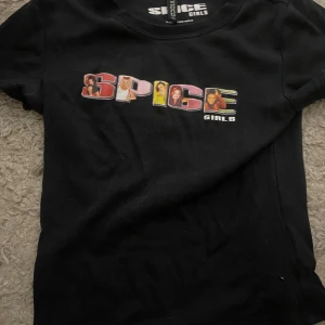 Svart Spice Girls t-shirt - Svart t-shirt med färgglatt Spice Girls-tryck på bröstet där bokstäverna är fyllda med bilder på medlemmarna. Klassisk passform och rund halsringning. 